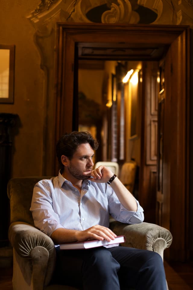 Davide Massiglia sitting in the room holding a red notebook and looking in the distance